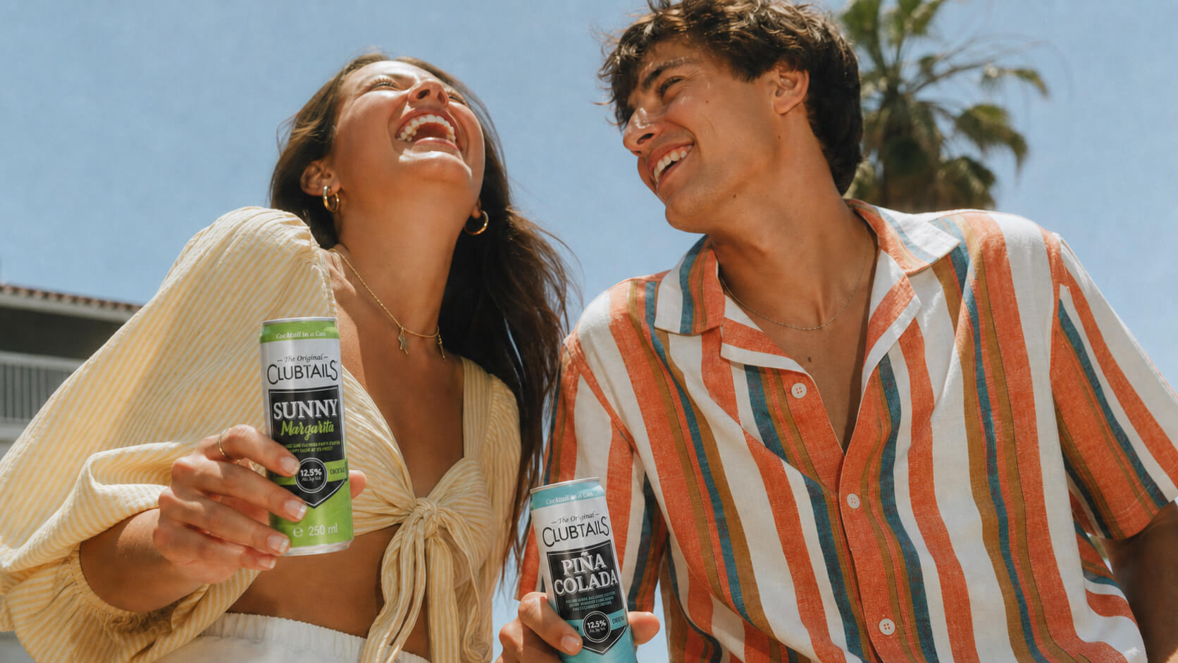 Two friends holding Clubtails Sunny Margarita and Piña Colada cocktail cans at a sunny poolside gathering.