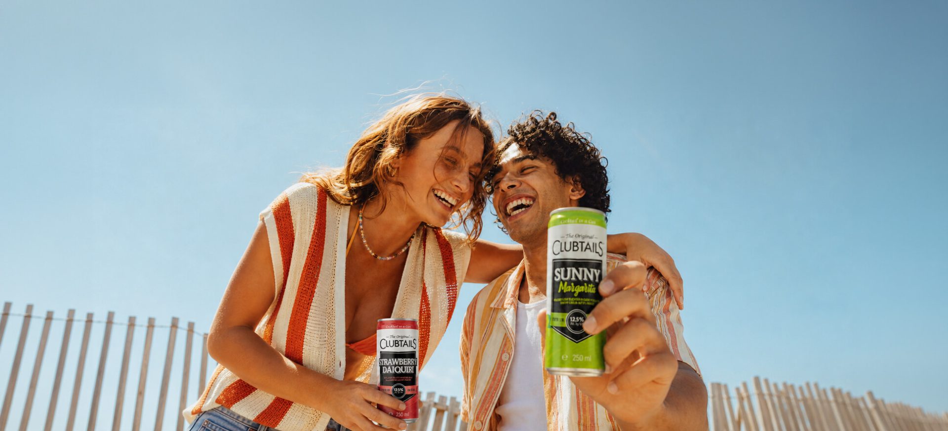 Two friends laughing at the beach while holding Clubtails Strawberry Daiquiri and Sunny Margarita cocktail cans.